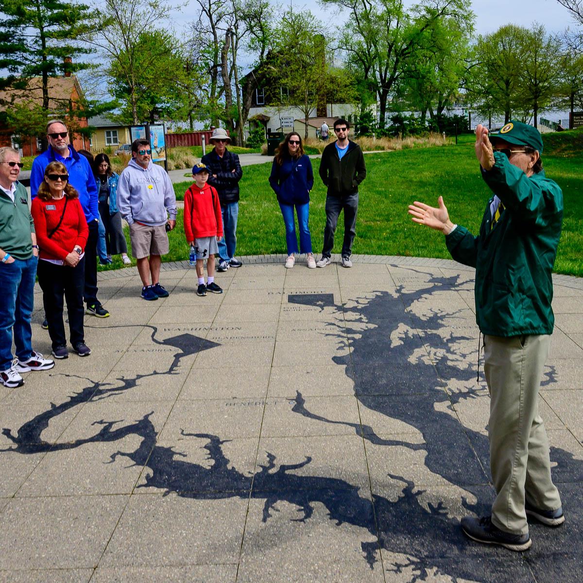 NPS volunteer in a green hat and jacket speaking to a group of visitors on a painted map outdoors.