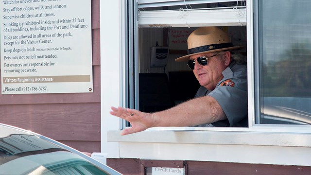 A park ranger, in a window, waving to a car. 