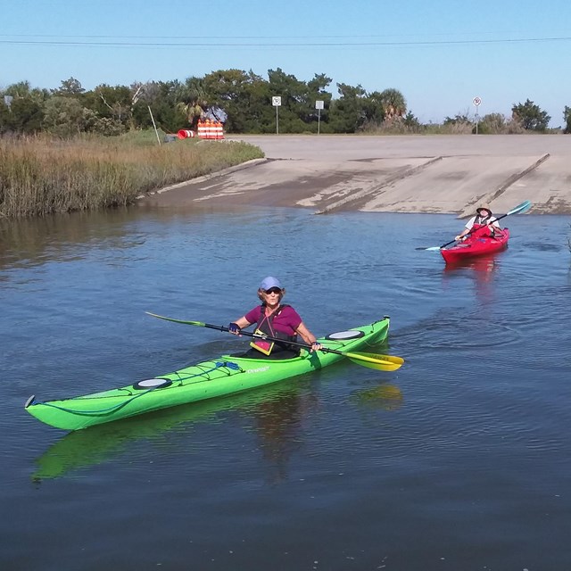 Two kayakers and kayaks pull away from a concrete boat ramp. 