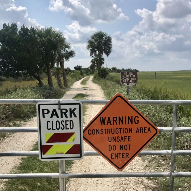 Gate with warning and park closed sign, dirt trail with trees and marsh in background