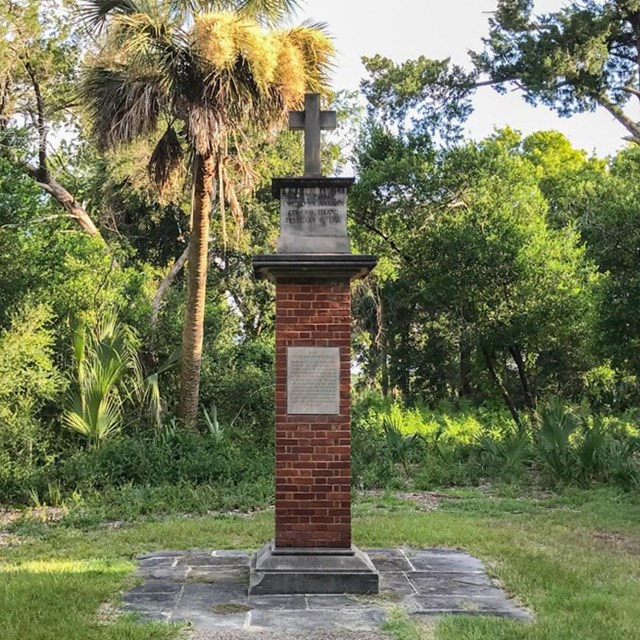 brick and concrete column monument with cross on top surrounded by trees and vegetation 
