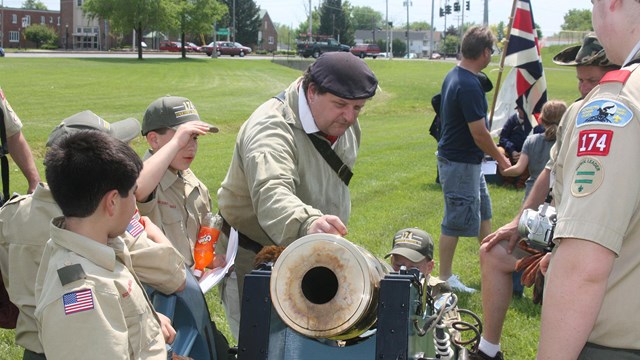 A Continental soldier stands over a brass cannon describing it to scouts. 