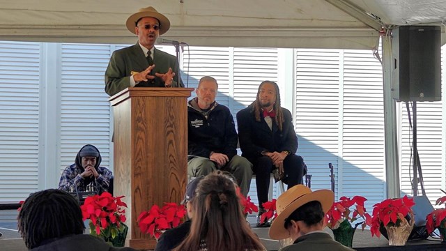 A man stands at a podium with two people sitting behind him. A group of people is seated facing him.