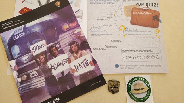 An open book lays on a desk. A closed copy lays on top. Below are a metal badge and sticker.