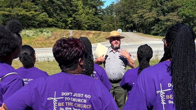 A man in a hat to a group of people in a grassy field.