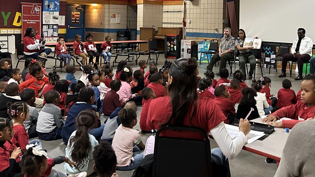 A group of about 100 children and teachers listen to rangers reading from a book. 