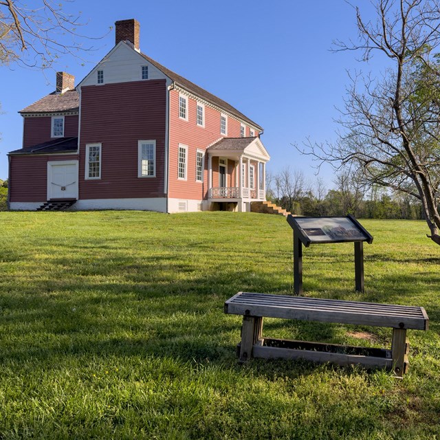 Two interpretive signs in front of a red two story farm house on a field of mowed green grass.