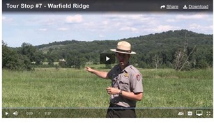 A park ranger points across a green field to two hills in the background. 