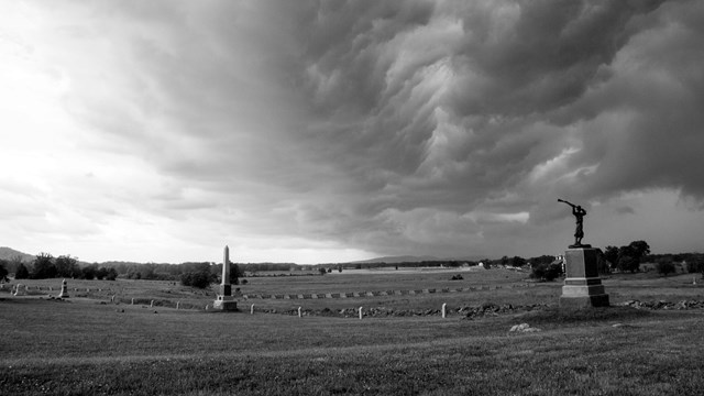 A black and white photo of storm clouds gathering over a field with monuments in the foreground.