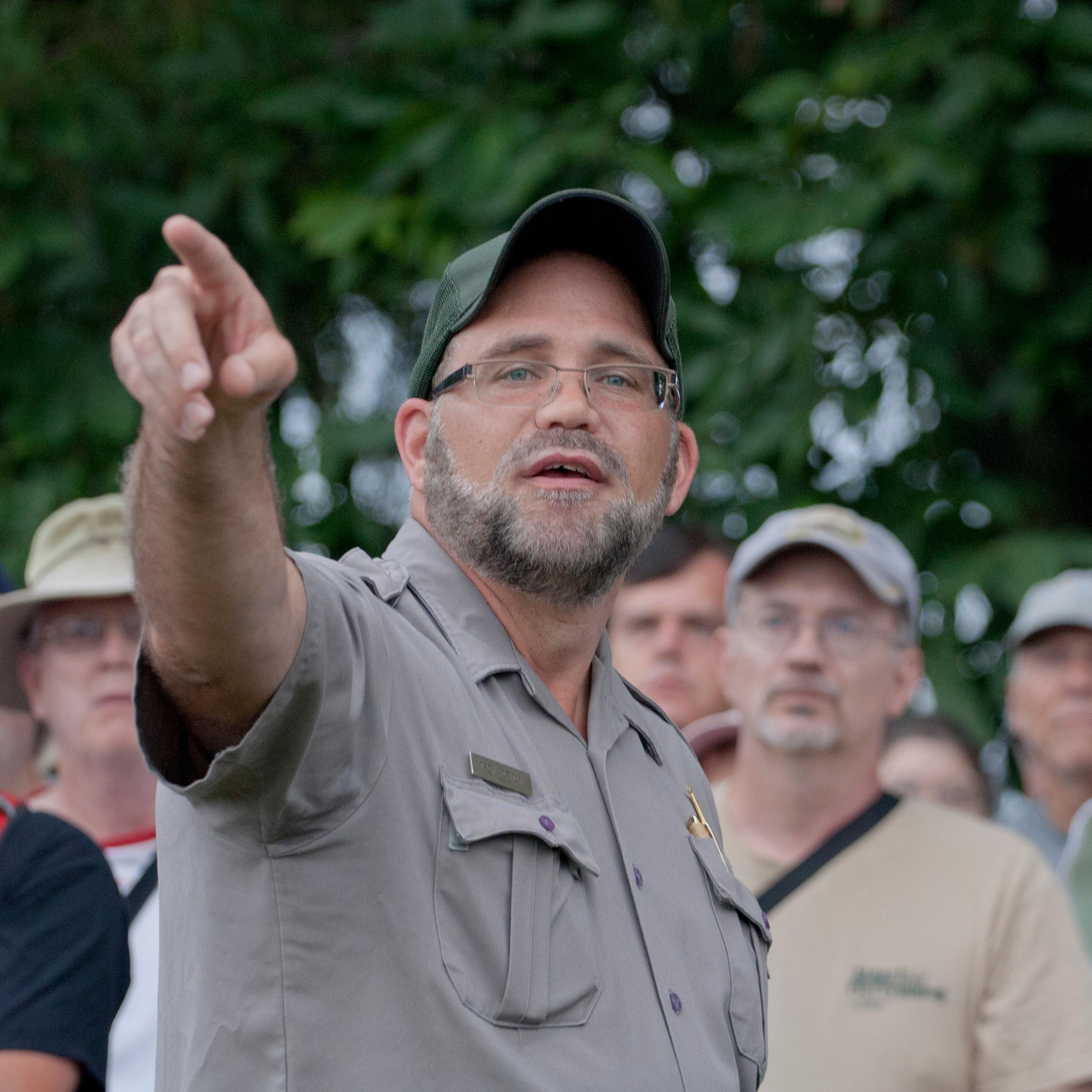A park ranger points out a battlefield location off in the distance while a large crowd of looks on.