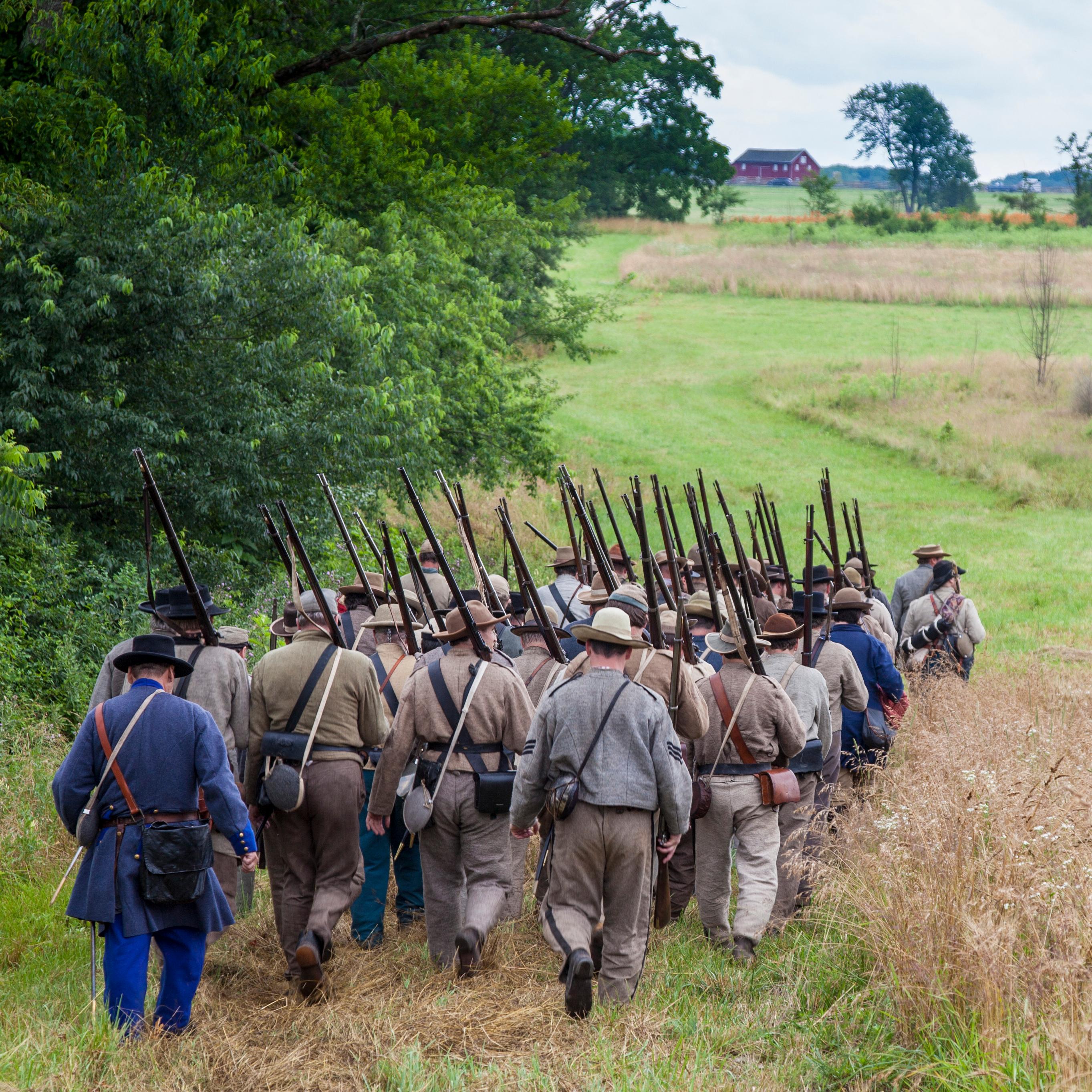 A group of Confederate living historians march across the field. A red barn is in the distance.