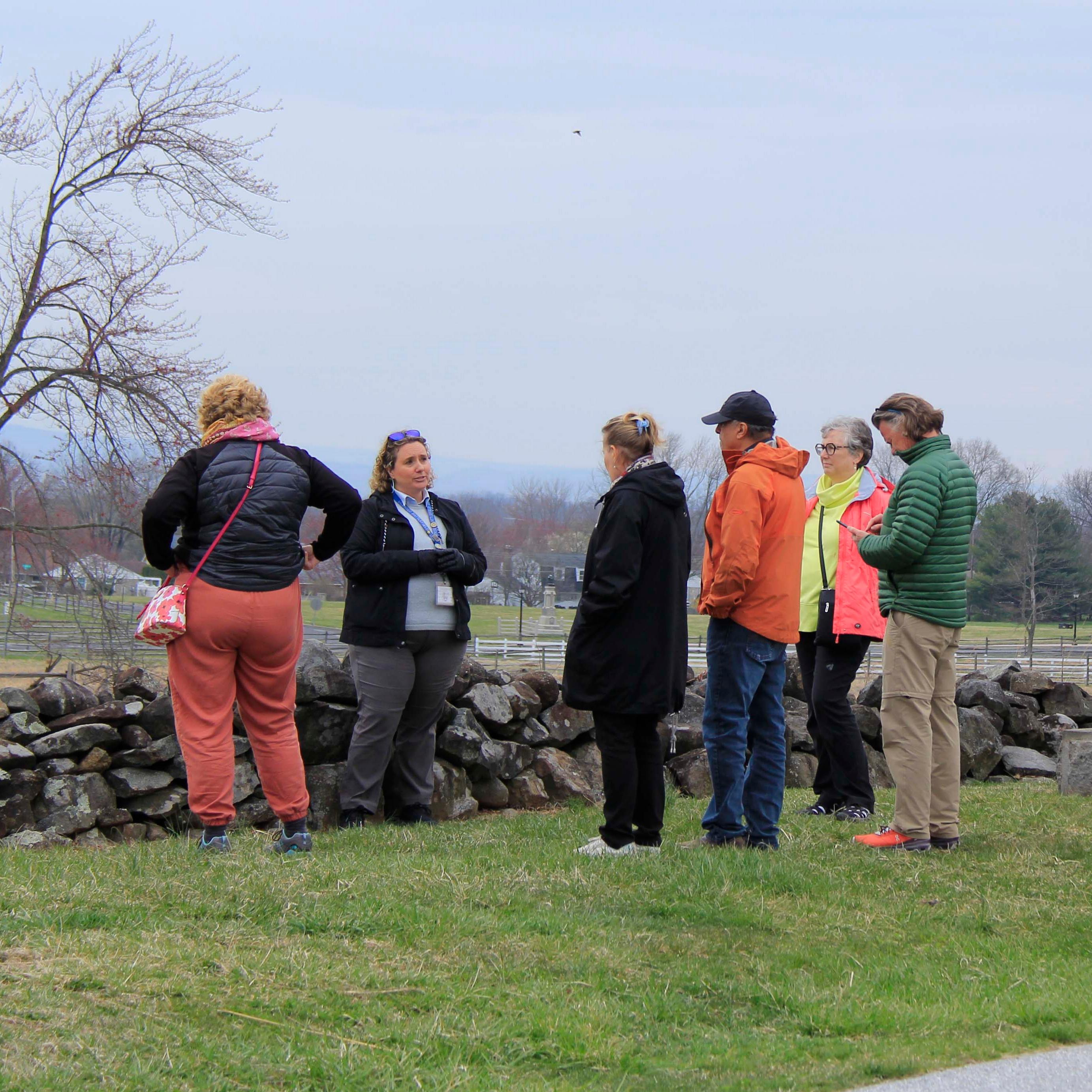 A group of visitors listen to a tour guide. The group stands in the grass next to a stone wall.