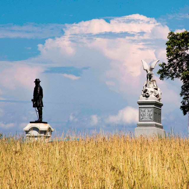A wheat field is in the foreground and two monuments are silhouetted against a blue sky and clouds.