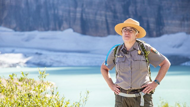 A park ranger stands in front of a blue lake and a mountain glacier. 