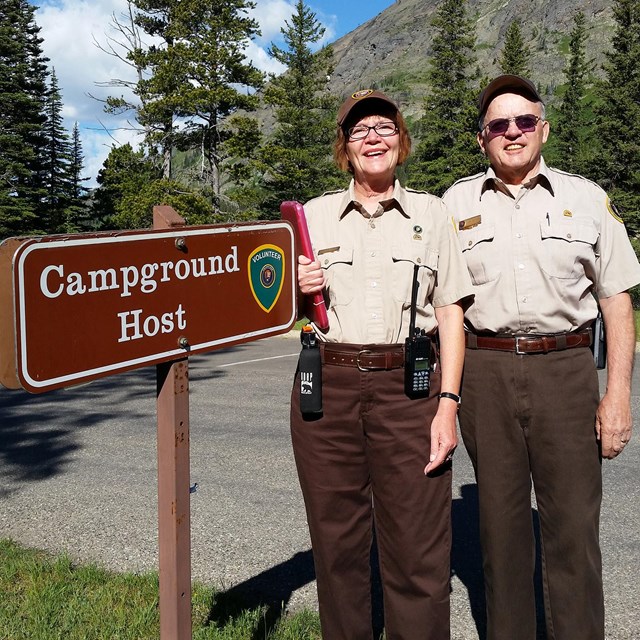 Two smiling, uniformed volunteers stand next to Campground Hosts sign