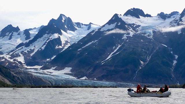 Large raft, with four people and gear, on lake in front of glacier.