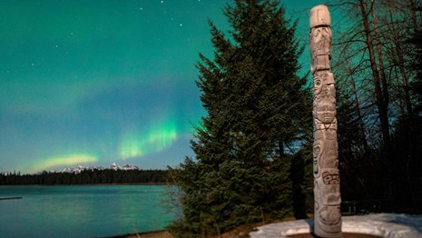 A totem pole rises high in front of spruce trees. Aurora and mountains in background.