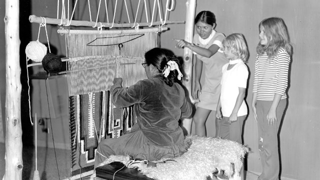 Historic photo of Navajo weaver at loom making a rug. Two children and a Park Ranger in a 1970s mini