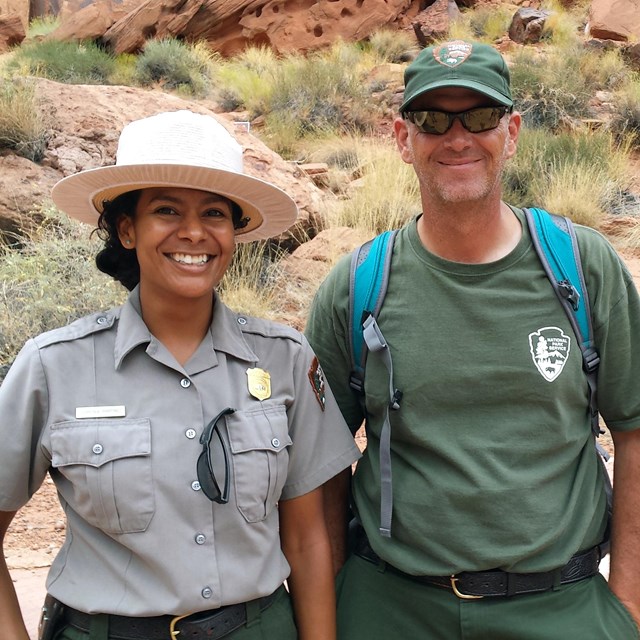 Two smiling rangers stand outside, one in formal uniform, the other in backcountry clothes