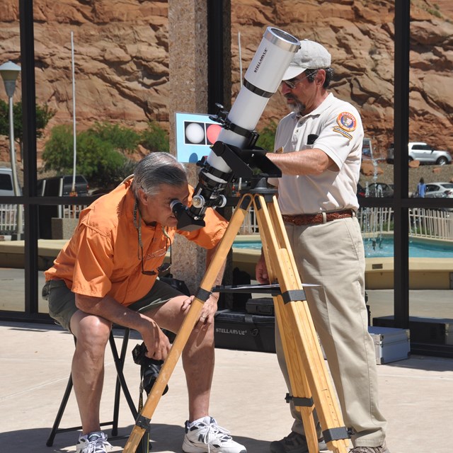 Man in volunteer uniform helps visitor peer through a solar telescope