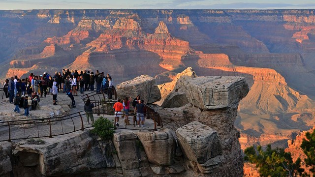 Crowd of visitors at a scenic overlook and watching orange light of sunset falling on Grand Canyon