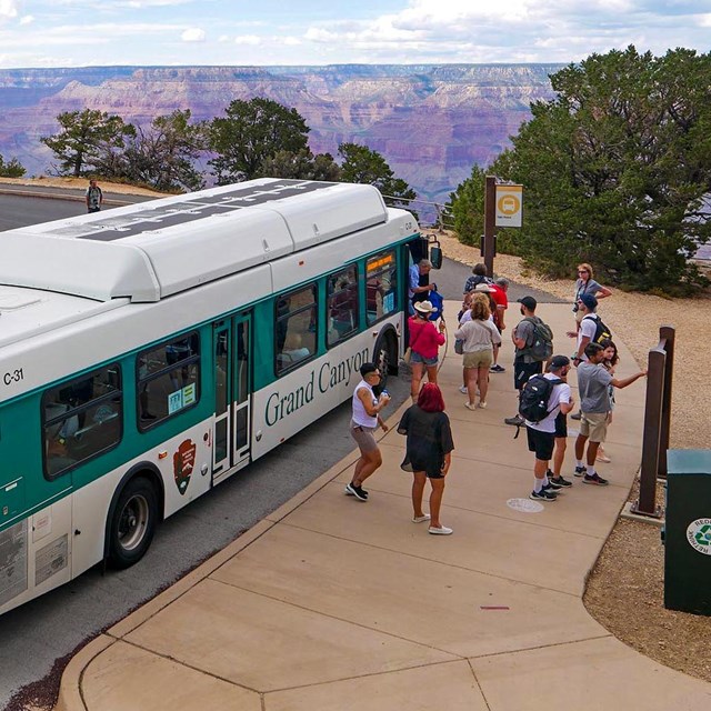 a green and white bus with passengers at a bus stop with a vast canyon landscape in the distance.