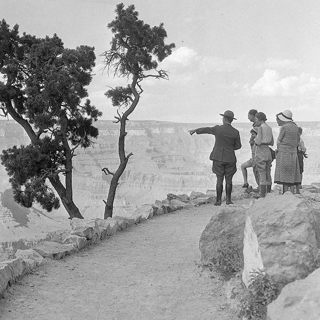 black and white photo from 1930's of park ranger escorting well dressed visitors on rim trail.