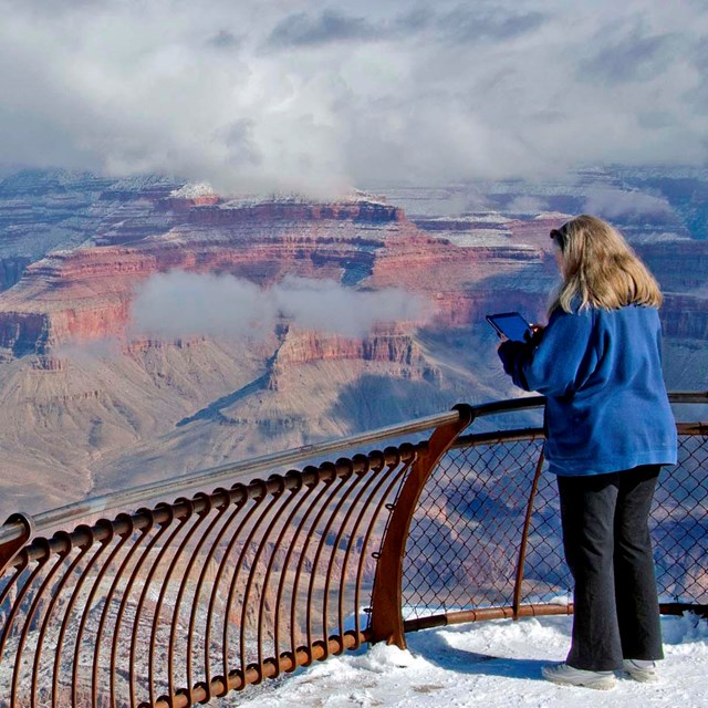 A woman behind a guardrail at a scenic overlook is watching winter clouds within a vast canyon 