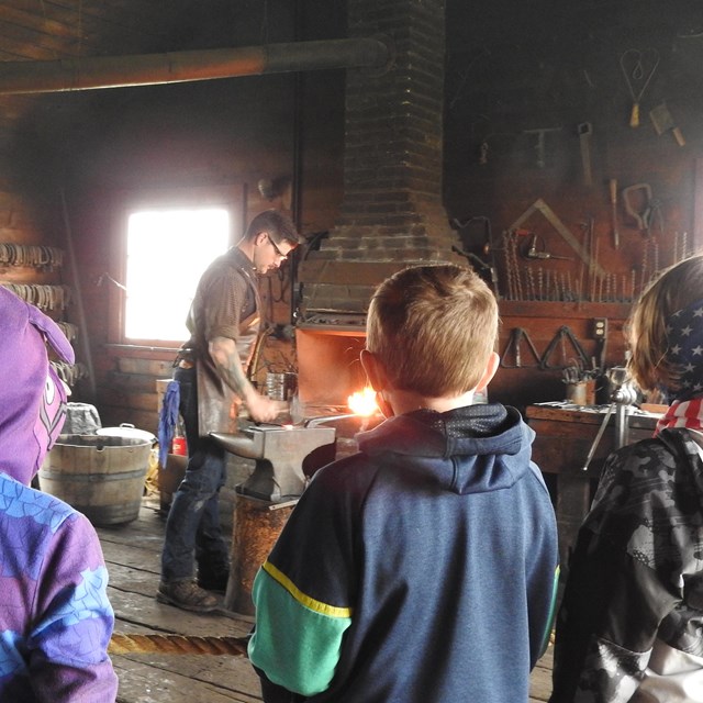 Group of students watches a ranger in historic dress as he hammers metal a the anvil, fire in forge 