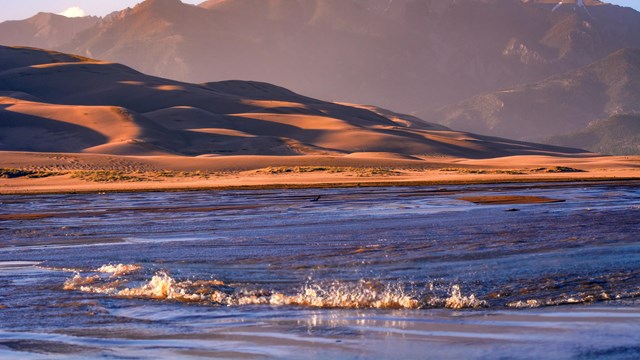 A shallow creek with a wave in front of dunes and a mountain