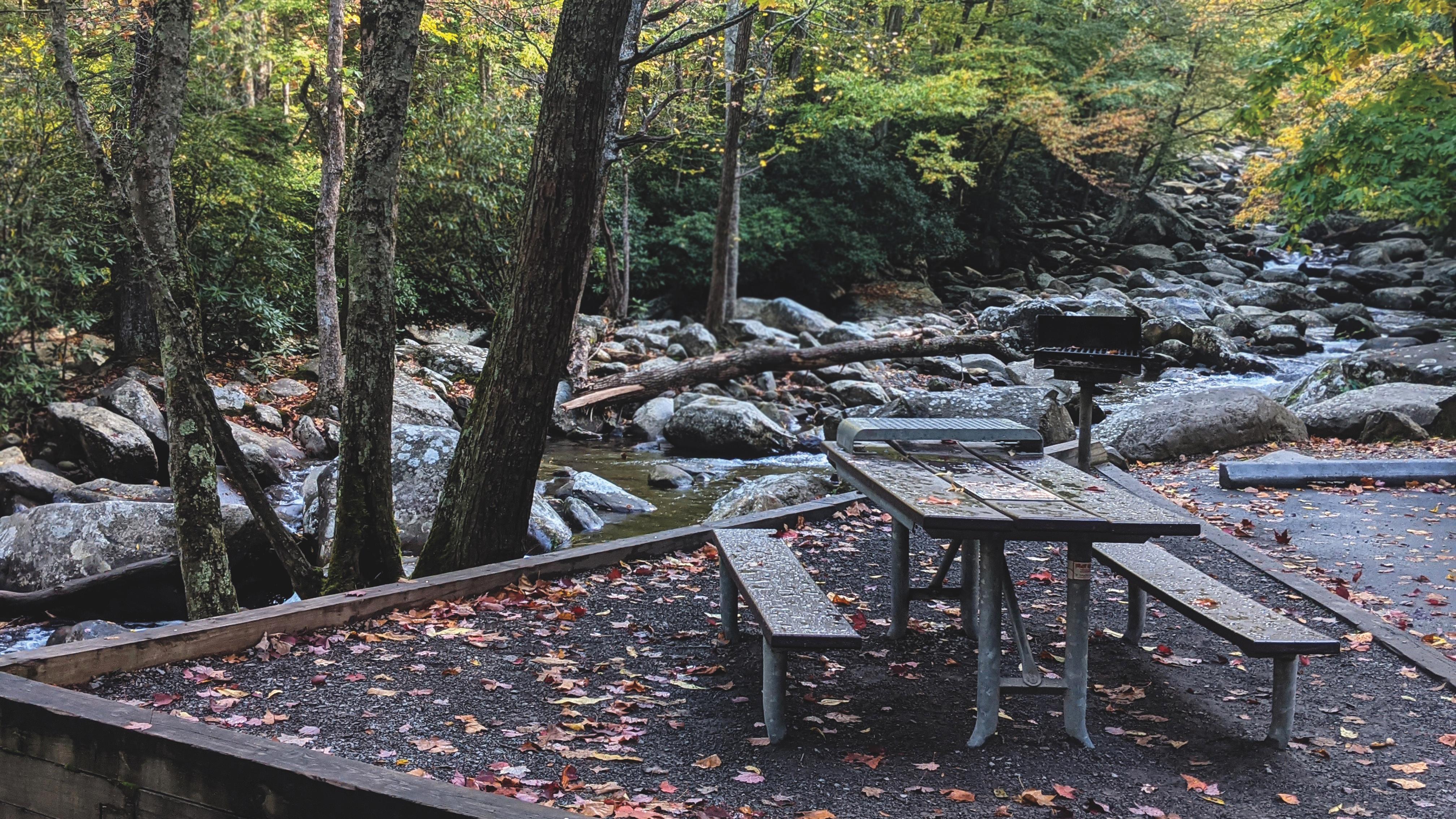 A wooden picnic table beside a river with large rocks and trees lining the sides