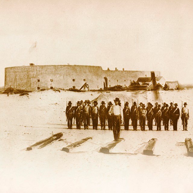 Historic image of soldiers standing in the foreground with a large masonry fort in the background.