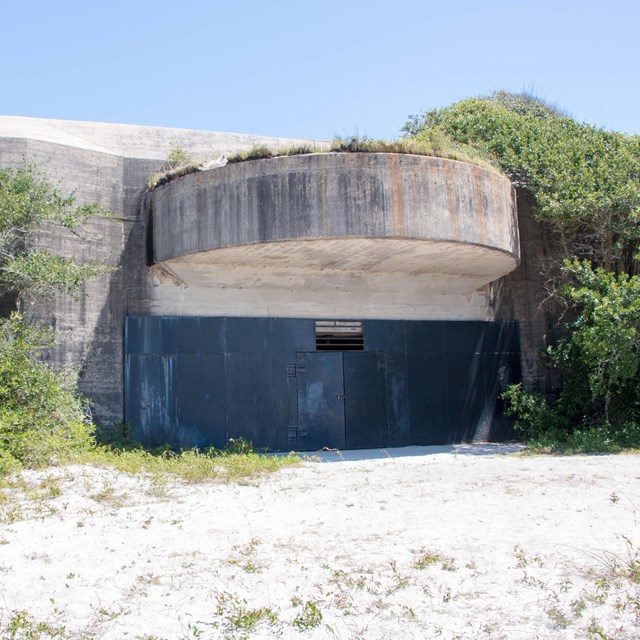 A massive concrete structure surrounded by vegetation sits on a beach.