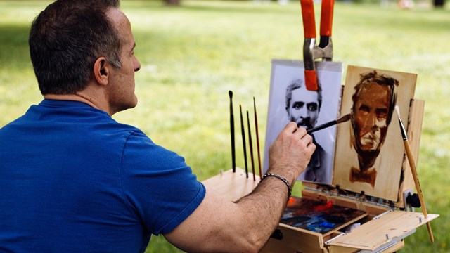 A man outdoors painting a portrait of George Washington Carver. 