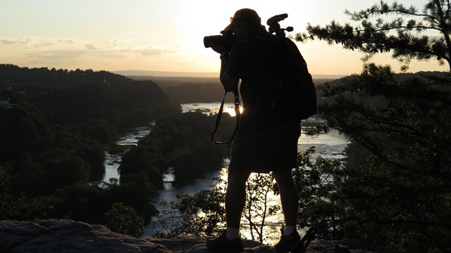 silhouette of a man taking a photograph; backdrop is trees, sky at sunset, and the river below 