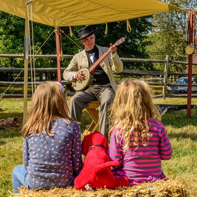 A park ranger in living history clothing playing a banjo for visitors. 