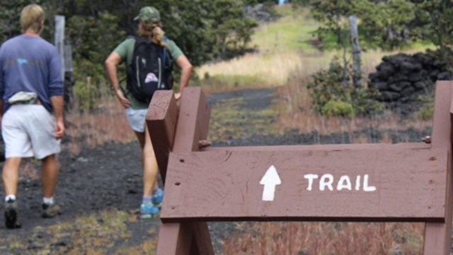 Two people walking along a trail with a large sign that says trail and a front facing arrow. 