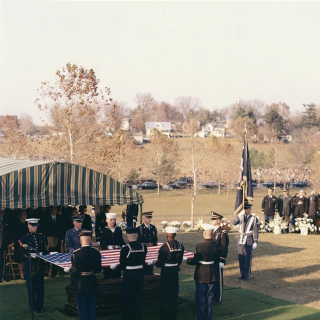 A 1960s color photo depicts a military honor guard burying a casket.