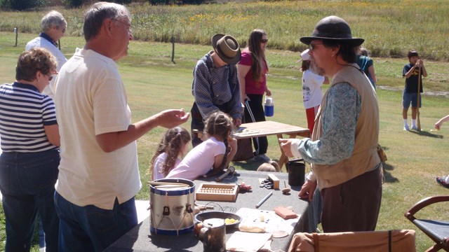 Man in period clothing speaks to visitors. On his table, an array of homesteading era toys and tools
