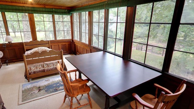 A sunny room with large windows looks out to trees. Inside, there is a long table and chairs.