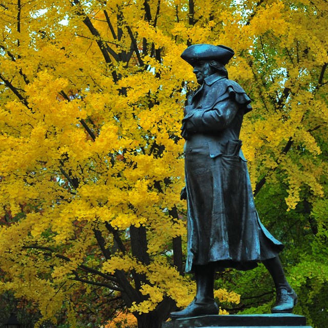 Color photo of a statue of a man in 18th century garb with fall foliage in the background.