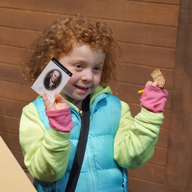 Color photo of a young girl holding up a Junior Ranger badge and a trading card.