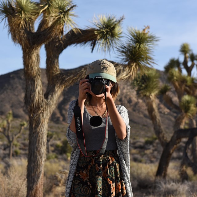 A young woman holds a DSLR camera to her face.