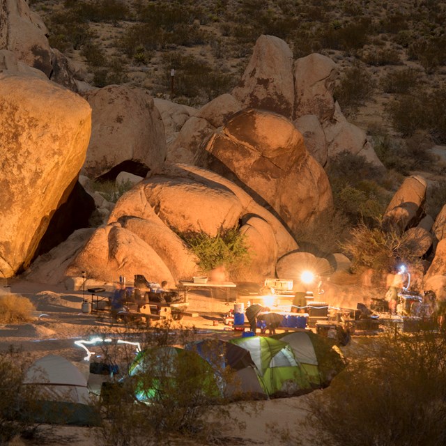 Color photo of a campsite at dark with a fire lit and headlamps leaving light trails.