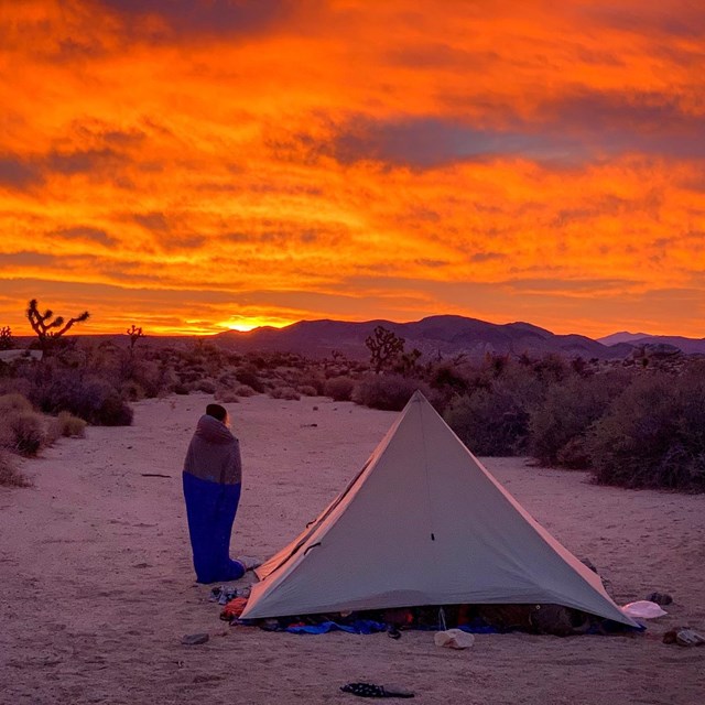 Person standing in a sleeping bag next to a tent and watching sunrise