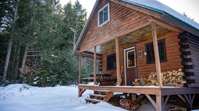 A brown wooden hut sits in an evergreen forest surrounded by snow. Deep green trees face the hut.