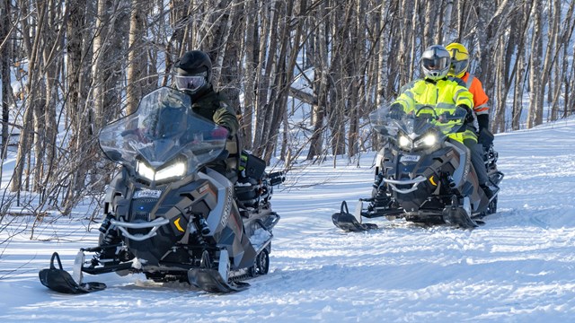 Two people on snowmobiles head down a snow packed trail lined with trees. 