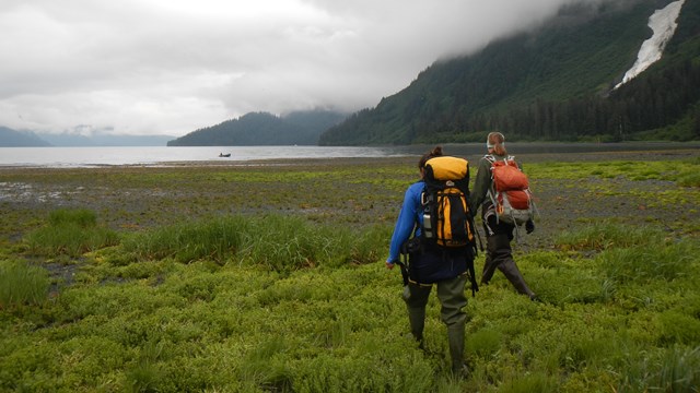 Two people with backpacks walk towards a small boat in a bay.