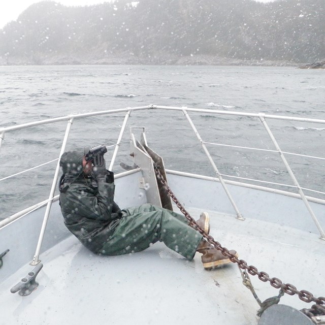 A person in rain gear looks through binoculars on a boat bow on a rainy day.