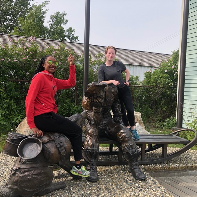 Two women sit on a bronze statue of a man seated on a sled and his dog
